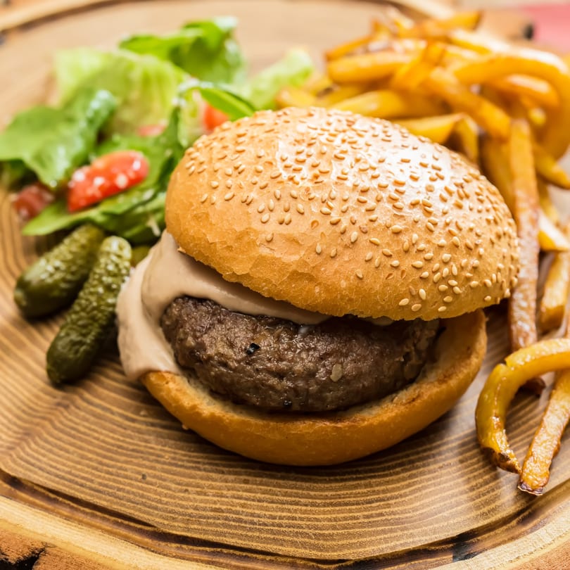 Zaab Burger with a sesame bun, fries, salad, and pickles on a wooden board.