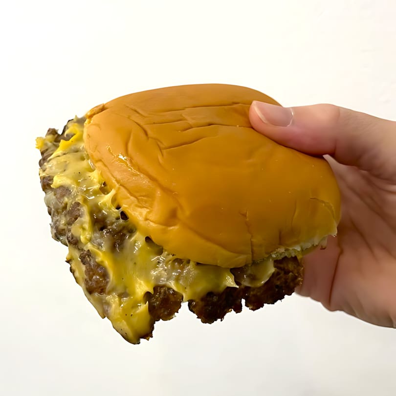 Hand holding a Zaab Burger with melted cheese dripping from two beef patties on a white background.